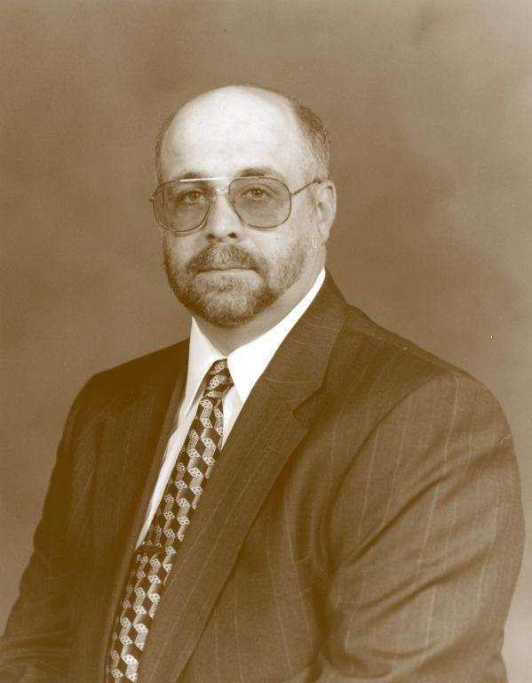 A man with a beard and glasses is wearing a suit and patterned tie, looking towards the camera.