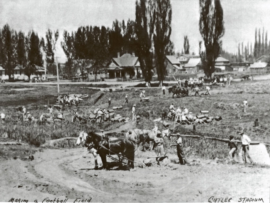 A group of people working in a field with horses pulling equipment. The scene is busy with individuals involved in various tasks, likely preparing the ground. In the background, there are trees and several houses. Handwritten text at the bottom reads "Making a Football Field" and "Cutler Stadium."