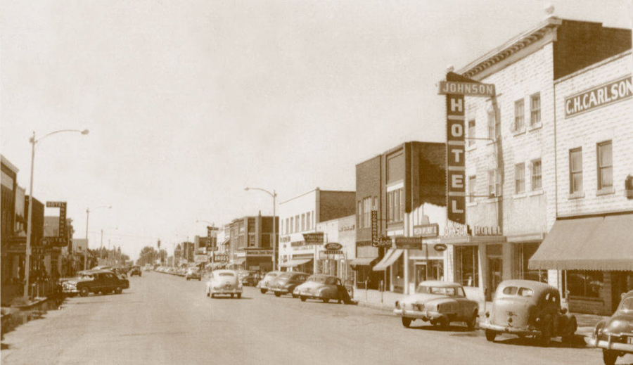 A street lined with buildings, featuring several parked cars. Signs visible on buildings include "Johnson Hotel" and "C.H. Carlson." Businesses have awnings, and street lamps are present. Traffic is minimal with a few cars on the road.