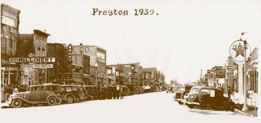 A busy street scene from 1939 in Preston. On the left, a row of buildings with signs including "Millinery" and "Rooms" is visible. A crowd of people is gathered along the sidewalk. Several vintage cars are parked along both sides of the street. On the right, there is a Texaco sign near a Chevrolet dealership and a cafe. The street features various storefronts and signs extending into the distance.
