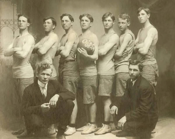 Seven young men stand in a line, all dressed in sleeveless shirts and shorts, likely a sports team. One holds a basketball labeled "O.S. 08-09." Two men in suits sit in front of the group. They are arranged in a studio setting with an ornate architectural backdrop.