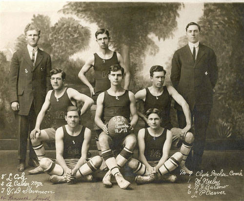 A group of seven men are posed in two rows. The front row, seated on the ground, consists of four men dressed in basketball uniforms. One holds a basketball with "OSA Champs, 1913" written on it. The back row includes three men, two in basketball uniforms and one in a suit. Another man in a suit stands at the far right. There is handwritten text at the bottom with names and the designation "Coach."