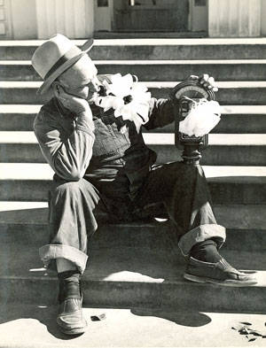 A person wearing a hat and overalls is sitting on steps with a thoughtful expression, resting their chin on their hand. They are holding a barometer adorned with large flowers. Scattered keys are visible on the step beside them.