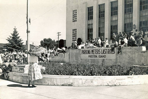 A crowd of people is gathered outside a large building with several windows. A woman with an umbrella stands in the foreground. There is a banner that reads "PARKING METERS LAST RITES PRESTON, IDAHO" on a raised stone platform where people are seated. A tree and a utility pole are visible in the background.