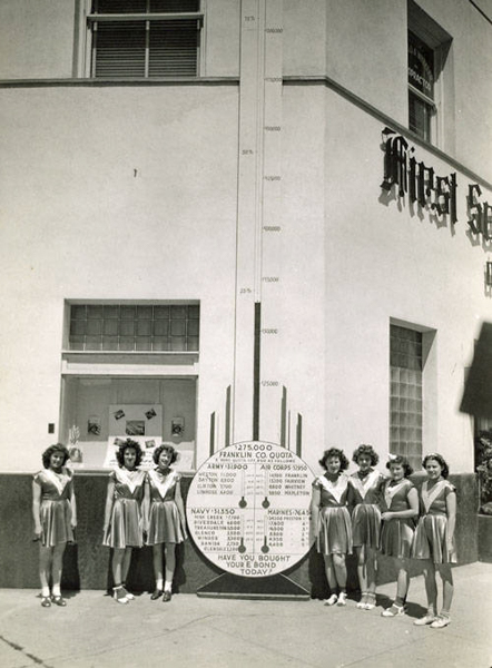 Seven women in matching outfits stand in front of a building. They are positioned beside a vertical sign with numbers and labels related to different military branches: Army, Navy, Air Corps, and Marines, with respective monetary goals. At the bottom, it reads "FRANKLIN CO. QUOTA" and asks, "HAVE YOU BOUGHT YOUR BOND TODAY?" The sign tracks financial contributions toward a quota of $275,000. The building has a window with some objects displayed and a partial sign with the word "Music" visible.