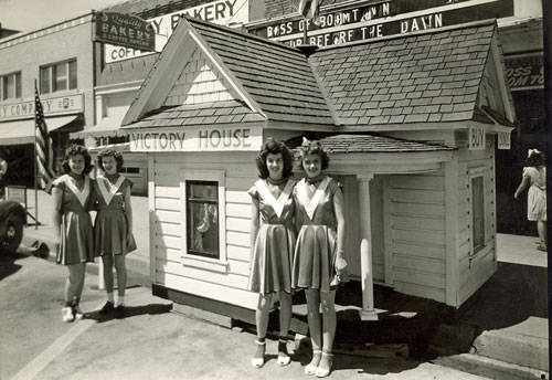 Four women in matching uniforms stand near a small house structure labeled "Victory House" on a street. In the background, there are additional buildings with signage that reads "Boss of Boomtown" and "Row Before the Dawn." An American flag is visible next to the women on the left.