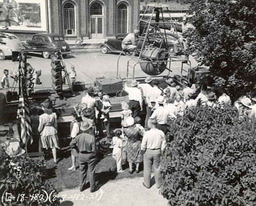 A crowd of people gathered around a platform featuring a spinning contraption suspended above it. Several adults and children stand nearby, some wearing hats. Across the street, vintage cars are parked in front of a large building with tall arched windows. A tree is in the foreground, and a portion of an American flag is visible. A billboard in the background reads "Enjoy Coca-Cola" with additional smaller text below.