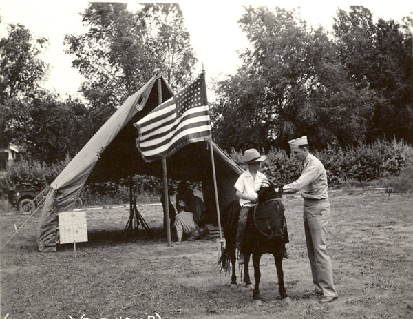A person is on horseback next to a man in a uniform. They are outside near a large tent displaying an American flag. There are people sitting under the tent. Trees and a vehicle are in the background.