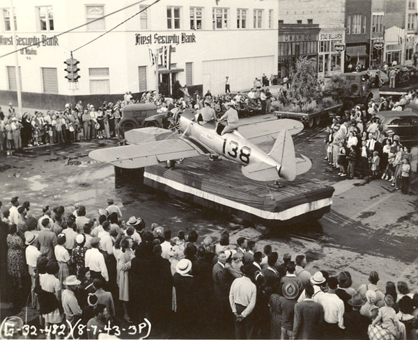 A crowd of people is gathered around a platform in the street, where a small aircraft with the number 138 is displayed. A person is seated in the cockpit of the aircraft. The adjacent building is marked First Security Bank. Numerous onlookers surround the scene, some dressed in hats. Urban buildings and a traffic light can be seen in the background, along with parked cars. Text on the image includes "G-32-482," "8-7-43-3P."