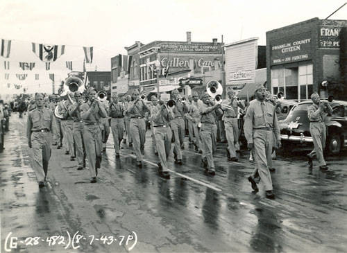 A group of marching band members in uniform walk down a wet street playing instruments, including tubas and trumpets. There are buildings lining the street with visible signage, such as "Fullen & Currey" and "Franklin County Citizen" with advertisements for printing and office supplies. A few cars are parked along the roadside and a crowd of onlookers stands nearby. Handwritten text at the bottom reads "(G-28-492) (8-7-43 7P)."