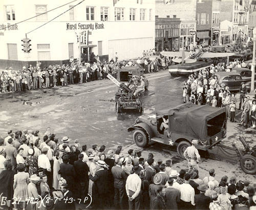 A large crowd of people is gathered along the sidewalks of a street, watching a parade. In the center, a military-style vehicle is towing equipment, possibly an artillery piece. The crowd includes men, women, and children, some wearing hats. Buildings line the street, including one labeled "First Security Bank." Another sign reads "Rexall Drugs." The street is wet, as if it has rained.