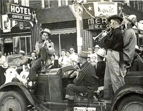 A group of musicians is performing on the back of a vintage truck. One man is sitting and playing a trumpet, while another is standing and playing a cornet. The truck is decorated, and a sign reads “Merchants Vets.” In the background, there are signs for a “Hotel Coffee Shop” and “Merck's Ice Cream.” A crowd of people watches the performance.