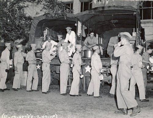 A group of uniformed soldiers is standing in a line beside a military truck. Some soldiers in the truck are serving food to those in line. Several soldiers are holding mess kits. The scene takes place outdoors near a building with windows. There are trees in the background, and the soldiers are wearing caps. The date "6-7-43" is visible at the bottom.
