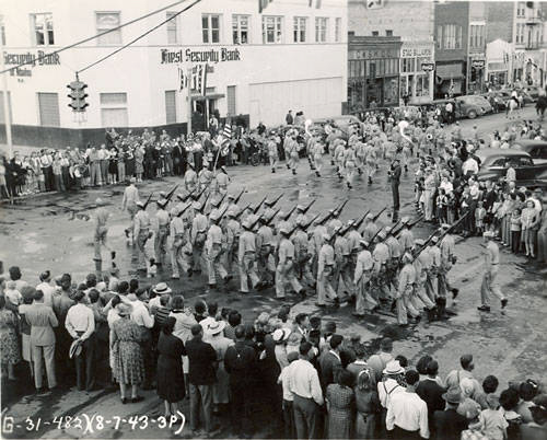 A large group of soldiers is marching in formation down a street, carrying rifles on their shoulders. A crowd of people lines both sides of the street, watching the parade. A building in the background displays the sign "Royal Security Bank." Several cars and more people are visible further down the street. Traffic lights hang above the intersection. The bottom of the image has handwritten text that includes the sequence "G-31-482 (8-7-43-3P)."