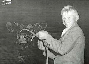 A young boy smiling while holding a rope attached to a cow. The boy is wearing a jacket and standing beside the cow, which has a harness on its head. There are no visible text elements in the image.