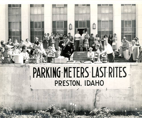 A group of people gathered on the steps of a building, some sitting and others standing. In front of the group, a sign reads "PARKING METERS LAST RITES PRESTON, IDAHO." A man appears to be speaking to the crowd, while others seem engaged in various activities, including serving from a table. The building features many windows in the background.