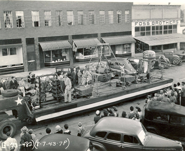 A street scene featuring a parade float displaying various machinery being viewed by a crowd. Several people are standing on the float, interacting with the equipment. The surrounding area has cars parked on the street and people watching the scene. A building in the background displays a sign reading "FOSS BROTHERS." Additional text includes a date and reference number: (8-7-43-7P) and [133-482].