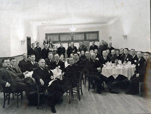 A group of men dressed in suits is seated and standing around tables in a room. The tables are set with cups and saucers. They are facing the camera. The room has light-colored walls with wall sconces and several closed doors or wooden panels on one side. There are no women present, and no text is visible.