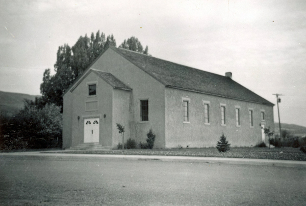 A rectangular building with a gabled roof sits next to a road. The facade features two doors with an arched design above them, and several tall windows line the side of the building. There are trees and small bushes around the building, and a utility pole is visible in the background.