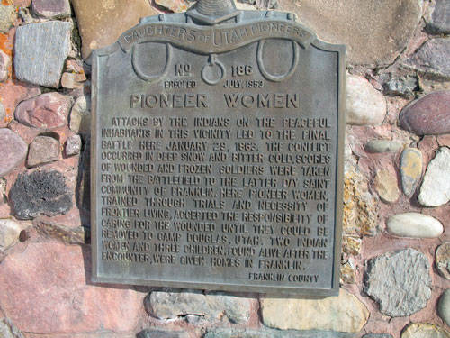 A plaque set into a stone wall is presented, featuring the emblem of the "Daughters of Utah Pioneers" at the top. It commemorates the "Pioneer Women" and describes attacks by Indians on peaceful inhabitants leading to a battle on January 29, 1863. The text tells of wounded and frozen soldiers being taken from the battlefield to the Latter-day Saint community of Franklin for care. Pioneer women managed hardships, providing aid and homes in Franklin County, as detailed in the dedication.