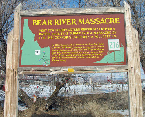 A wooden sign in a snowy landscape with leafless trees in the background. The sign reads "Bear River Massacre" at the top, followed by text detailing historical events. It describes a battle that turned into a massacre by Col. P.E. Connor’s California Volunteers, highlighting the small number of Northwestern Shoshoni survivors. The text mentions the date January 1863 and gives a brief account of the incident. There are two small number signs on the sides, showing "91" and "216."
