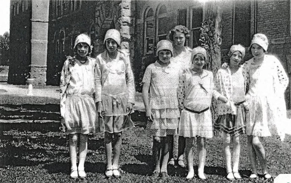 A group of eight young girls standing on grass in front of a large building. They are wearing dresses and cloche hats, typical of early 20th-century fashion. The building behind them has arched windows and stone pillars.