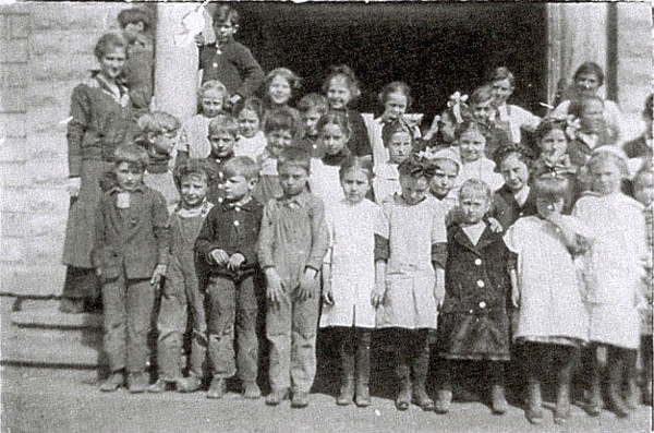 A group of children and a few adults standing together on the steps of a building. The children are dressed in various outfits, some with bows in their hair. They are positioned in several rows, with some older girls standing in the back. The building has a brick exterior and a large doorway.