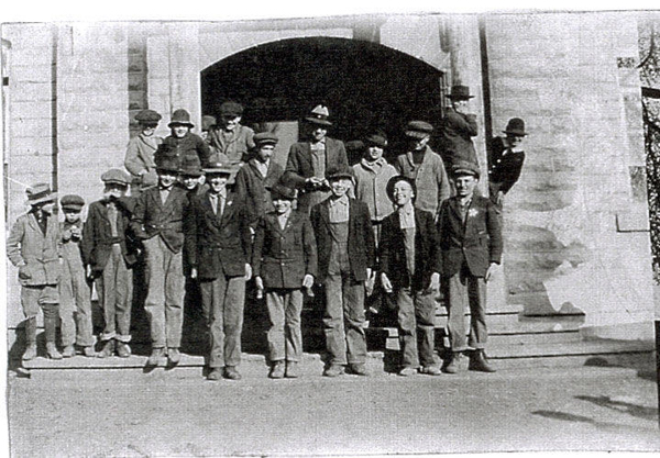A group of nineteen boys and young men standing on the steps of a large stone building. Most are wearing jackets and hats. The entrance of the building features an arched doorway, and there are other people visible on the steps and near the doorway.