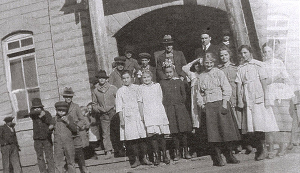 A group of children and adults standing in front of a large building with an arched entrance. The children, mostly girls, are wearing dresses, while the adults in the background have hats and coats. Some boys are wearing caps and jackets. There are windows visible on the side of the building.