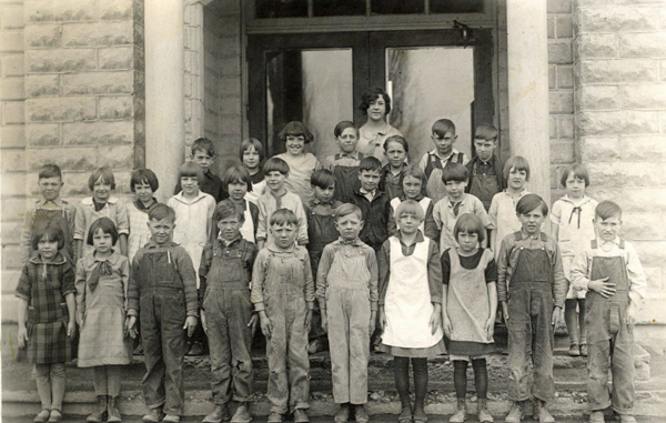 A group of children and a woman standing on the steps of a brick building. The children are dressed in various outfits, including dresses, overalls, and shirts. The woman stands behind the children near the building's entrance.