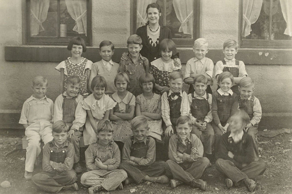 A group of young children, consisting of both boys and girls, posing in several rows in front of a building with large windows. An adult woman stands behind them, smiling. The children are wearing a mix of dresses and overalls. The atmosphere appears friendly and organized.