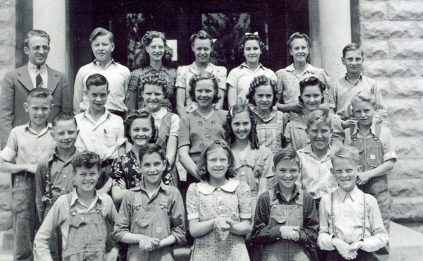 A group of children and an adult standing in front of a stone building. The group includes boys and girls, with the boys mostly wearing overalls or shirts, and the girls dressed in patterned dresses. The adult is wearing a suit and tie. The individuals are arranged in several rows, smiling at the camera.