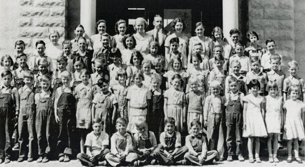A large group of children and a few adults pose for a group portrait in front of a building. The children are wearing a variety of clothes including overalls and dresses. The adults are standing in the back row. All are smiling or looking toward the camera.