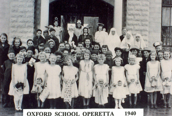 A group of children and a few adults stand in front of a building entrance. The children are wearing various costumes and holding dolls. Some adults are in costume as well, including outfits resembling nuns. The text at the bottom reads "OXFORD SCHOOL OPERETTA 1940."