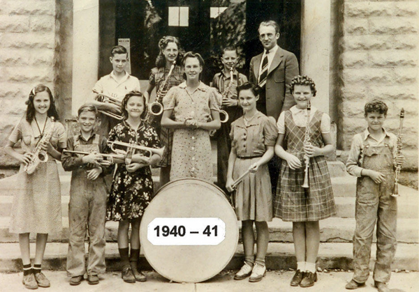 A group of young students and an adult stand on steps in front of a stone building. The children hold musical instruments, including saxophones, clarinets, a trumpet, a trombone, and a drum. A sign in front of the drum reads "1940 - 41." The adult, standing in the back, wears a suit and tie. The children are dressed in patterned dresses and overalls.