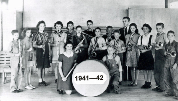 A group of young people is standing in a room, each holding a musical instrument such as clarinets, trumpets, and saxophones. At the front, a girl is kneeling beside a large drum that is labeled "1941—42." There are benches and a wooden structure in the background.