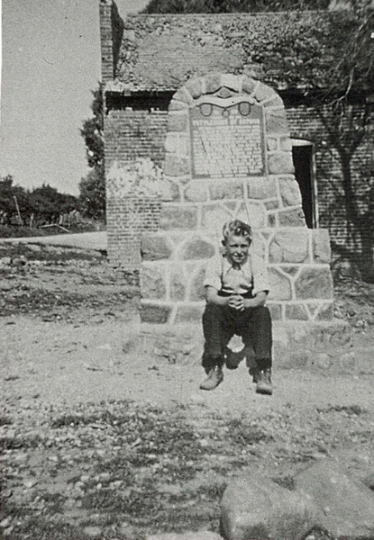A child sits on a stone structure, which has a plaque with some text on it. The child is wearing a light-colored shirt and dark pants. Behind the structure, there is a brick building. Trees are visible in the background.