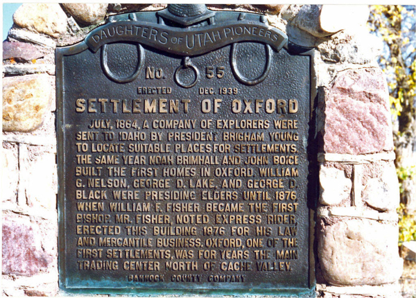 A metal plaque mounted on a stone wall with the inscription "Daughters of Utah Pioneers No. 55 Erected Dec. 1939." The plaque title reads "Settlement of Oxford." The text describes the history of Oxford, Idaho, starting in July 1864, when explorers sent by President Brigham Young established the settlement. It mentions Noah Brimhall and John Boice building the first homes, and William G. Nelson, George D. Lake, and George D. Black serving as presiding elders until 1876. William F. Fisher is noted as becoming the first bishop. The plaque commemorates Oxford as a major trading center in Cache Valley and mentions Fisher's involvement in erecting a building for his law and mercantile business. At the bottom, it states "Bannock County Company."