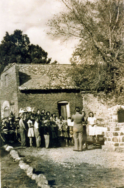 A group of people, including children, gathered in front of an old brick building with a slanted roof. A man stands in the center facing the crowd, appearing to address them. A woman is standing near a table with books placed on it. There is a stone structure to the right, and trees surround the area.
