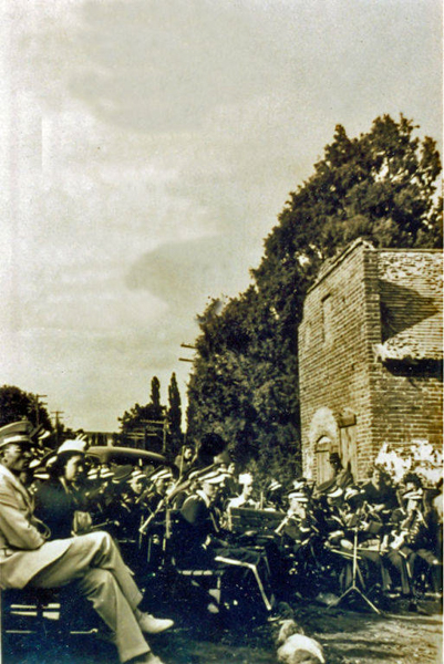 A group of people, likely a band, wearing uniforms and hats, are seated outside in rows with musical instruments. In the foreground, a few individuals are seated on chairs, seemingly listening. A brick building and some tall trees are in the background.