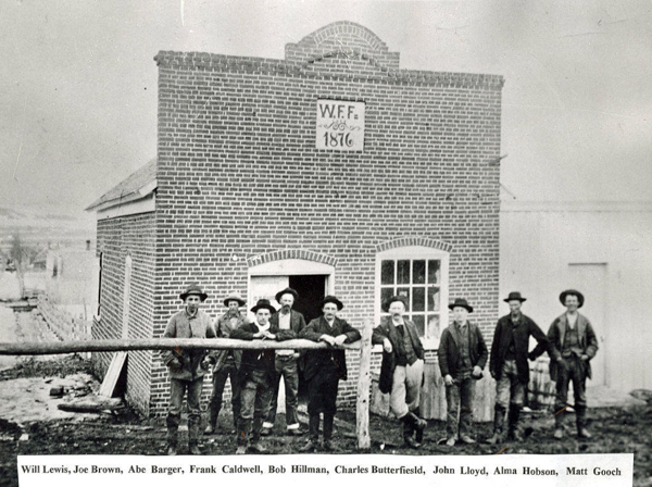 A group of nine men standing in front of a small brick building with arched windows and a sign on the facade that reads "W. F. F. 1876." The men are dressed in 19th-century attire, some wearing hats. Below the image, names listed include Will Lewis, Joe Brown, Abe Barger, Frank Caldwell, Bob Hillman, Charles Butterfield, John Lloyd, Alma Hobson, and Matt Gooch.