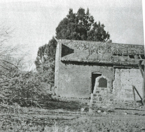An old, deteriorating building with a partially collapsed roof and a rough stone structure in front. The building is surrounded by overgrown vegetation and trees.