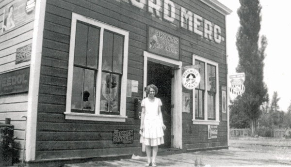 A woman stands in front of a wooden building with large windows and a door. The signage on the building includes "BROWN'S ICE CREAM," "CHICKEN DINNERS," and "BUY WHEAT FLAKES." Above the door, the words "RD MERC" are visible, along with various other smaller signs. Trees and a fence can be seen in the background.