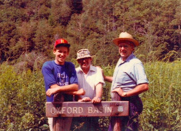 Three men standing behind a wooden sign that reads "Oxford Basin" in a lush, green forest setting. One man on the left wears a red cap and a shirt with partially visible text. The man in the middle is wearing a light-colored hat and a white shirt, while the man on the right is wearing a straw hat and a light blue shirt.