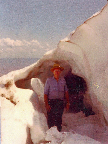 A person wearing a hat and a shirt stands inside a cave made of snow or ice. The cave has smooth, rounded edges and opens out to a landscape with a sky and distant clouds in the background.