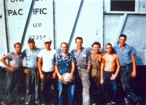 Eight men standing in front of a train car with the words "PACIFIC" and "U.P. 03350" visible. One man in the center is holding a ball. Some men are wearing work clothes, while others are in casual attire.