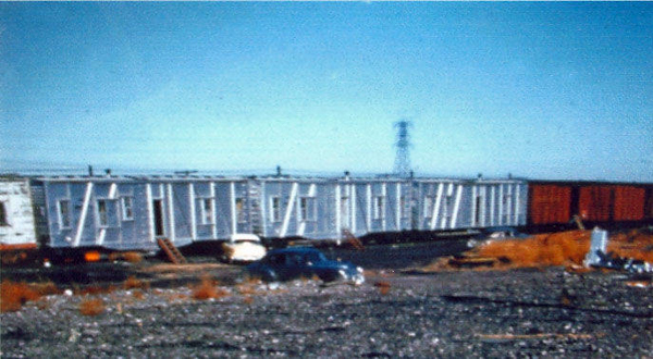 A freight train with several boxcars in a rail yard. There are two cars parked near the train, and a utility tower can be seen in the distance. The ground is covered in gravel and some scattered plants.