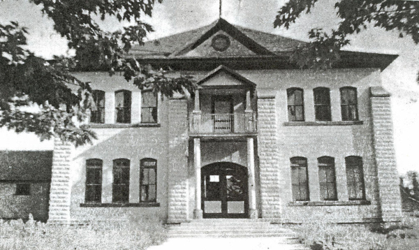 A two-story, symmetrical brick building is shown with a central entrance featuring a small porch and steps leading up to double doors. The building has multiple tall, narrow windows on both floors. Trees partially frame the view of the building.