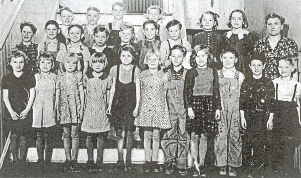 A group of children and a woman, possibly a teacher, stand in rows on a staircase. The children are wearing a variety of clothing styles, including dresses and overalls. The teacher is wearing a patterned dress. All are facing forward, smiling or with neutral expressions. There is no text visible in the image.