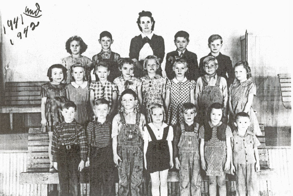 A group of children and one adult arranged in three rows, posing indoors. The adult stands in the center of the back row, while the children, wearing a variety of dresses and overalls, stand and sit on benches. Handwritten text in the upper left corner reads "1941" and "1942."